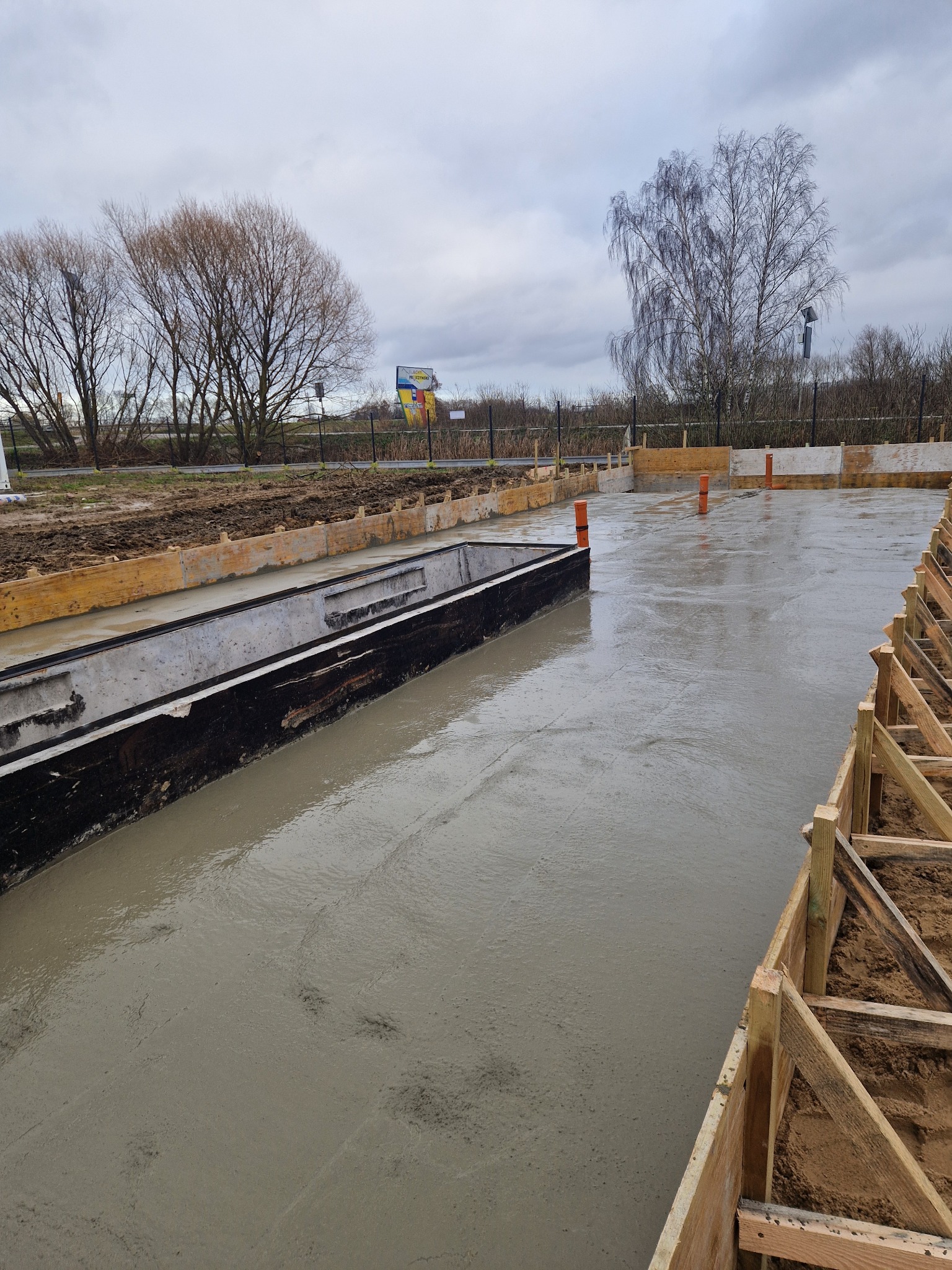 Zalany betonem fundament z drewnianym szalunkiem i wystającymi rurami wentylacyjnymi. W tle drzewa i pochmurne niebo. Widok z góry na plac budowy.