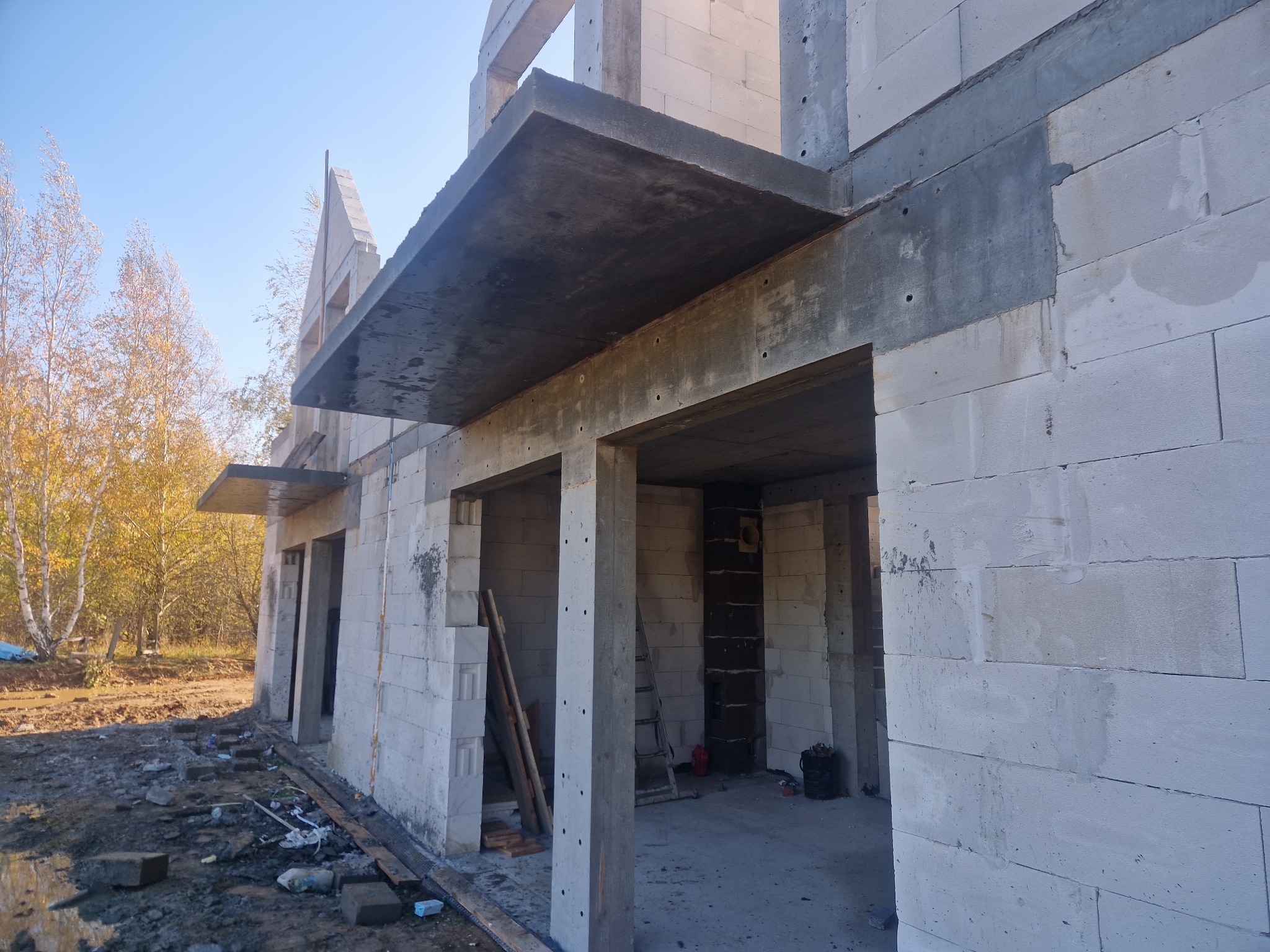 Surowy stan domu z betonu komórkowego w trakcie budowy, widoczne stropy i ściany, na zewnątrz drzewa z jesiennymi liśćmi, ziemia częściowo błotnista.