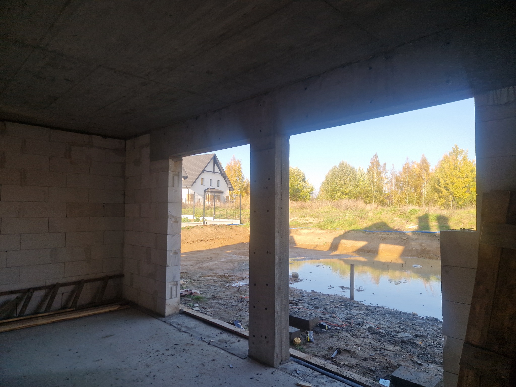 Surowy stan domu w budowie: ściany z betonu komórkowego, widok na staw i dom w tle przez otwór okienny. Na podłodze widoczne deski i drabina.