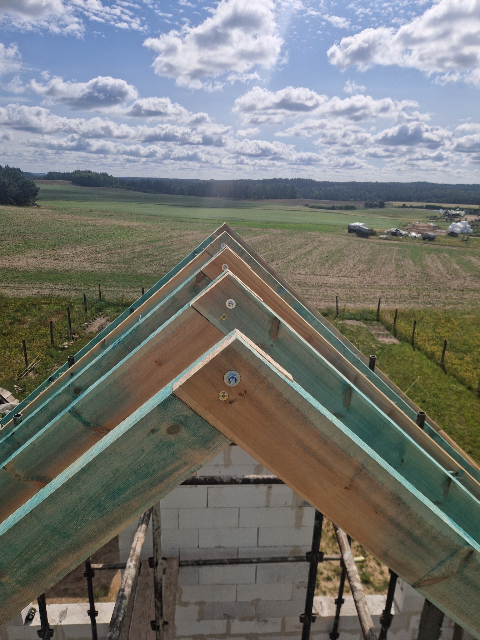Konstrukcja dachu z impregnowanego drewna na tle krajobrazu z polami i lasem, widok z góry, jasne niebo z chmurami. Elementy rusztowania widoczne w dolnej części kadru.