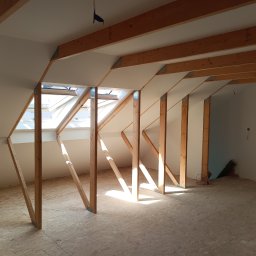 PIOTR DOMAŃSKI - Poddasze z drewnianą konstrukcją dachu i oknami połaciowymi, przygotowane do dalszych prac wykończeniowych. Widoczne drewniane belki i płyty OSB na podłodze.