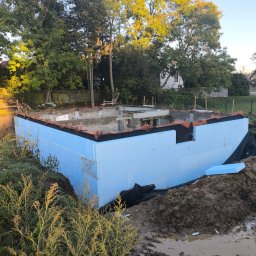 PIOTR DOMAŃSKI - Piwnica w trakcie budowy: fundamenty z izolacją termiczną z niebieskiego styropianu, widoczne zbrojenia i bloczki betonowe. Tło: drzewa i fragment budynku.