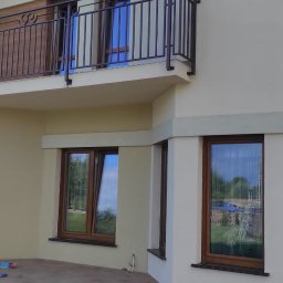 PIOTR DOMAŃSKI - Jasna elewacja domu jednorodzinnego z balkonem i brązowymi oknami. Widoczny fragment tarasu z kostki brukowej. Ujęcie z dołu, w tle błękitne niebo.