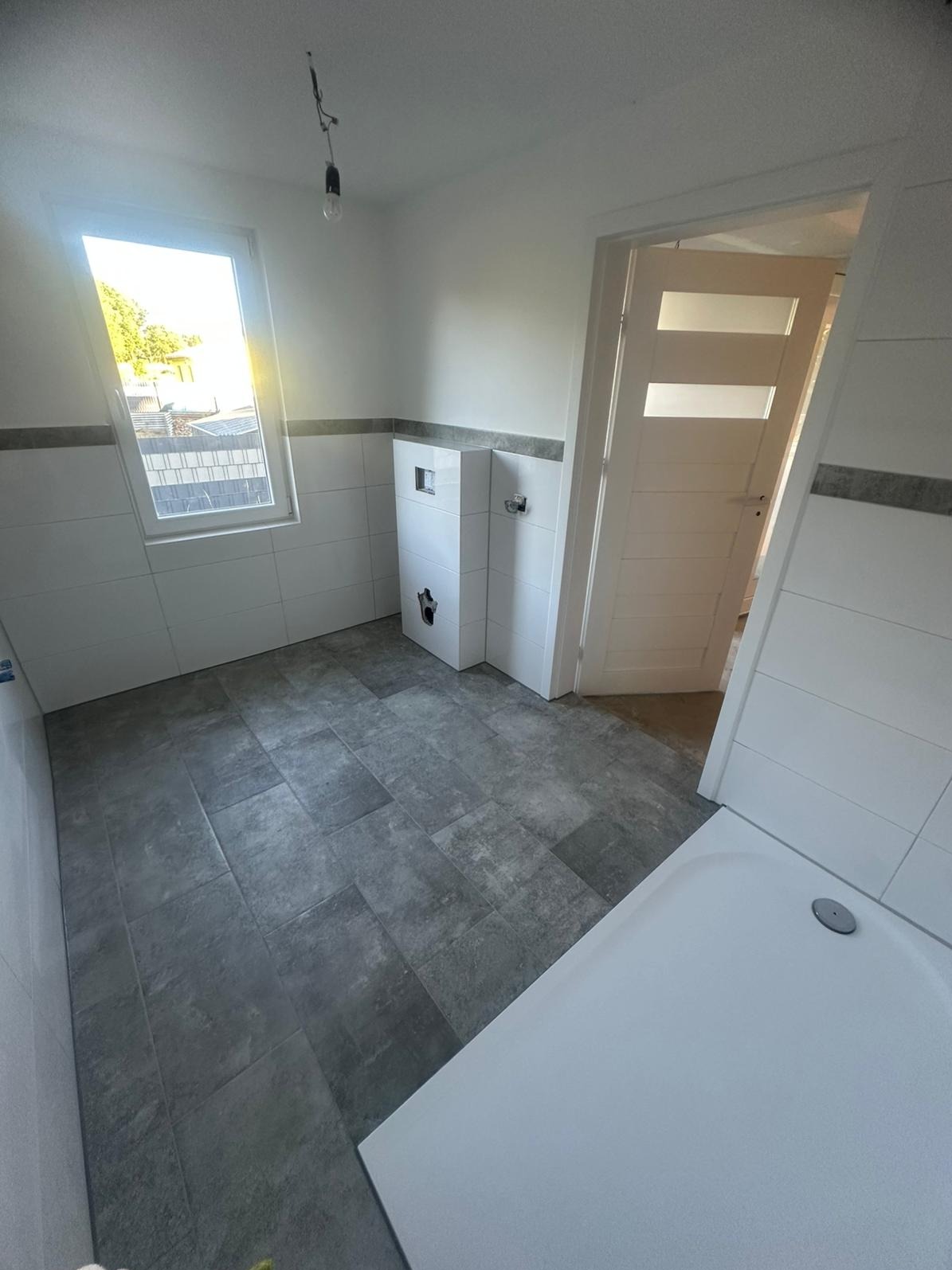 Wykończona łazienka z szarymi płytkami podłogowymi i białymi płytkami ściennymi. Widoczna kabina prysznicowa, okno i białe drzwi z poziomymi przeszkleniami.