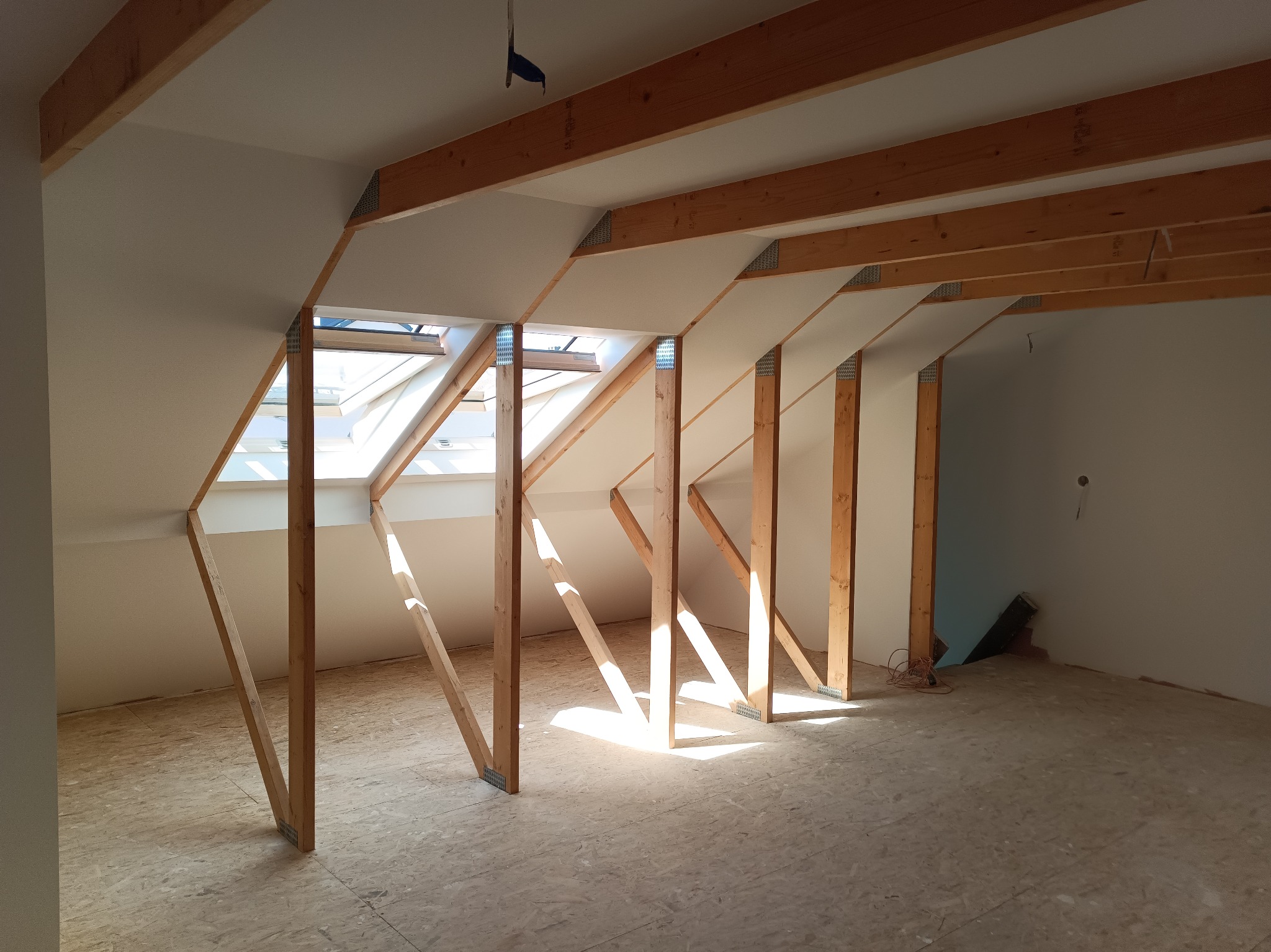 Poddasze z drewnianą konstrukcją dachu i oknami połaciowymi, przygotowane do dalszych prac wykończeniowych. Widoczne drewniane belki i płyty OSB na podłodze.