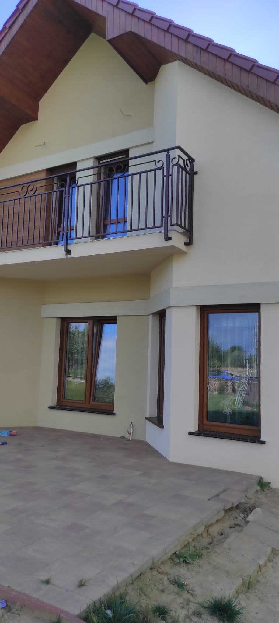 Jasna elewacja domu jednorodzinnego z balkonem i brązowymi oknami. Widoczny fragment tarasu z kostki brukowej. Ujęcie z dołu, w tle błękitne niebo.