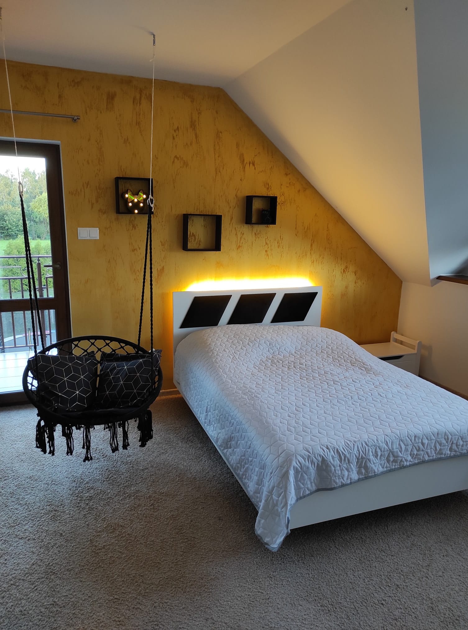 Żółta ściana w sypialni na poddaszu z wiszącym fotelem makrama, białym łóżkiem z podświetlanym zagłówkiem i dekoracyjnymi półkami.