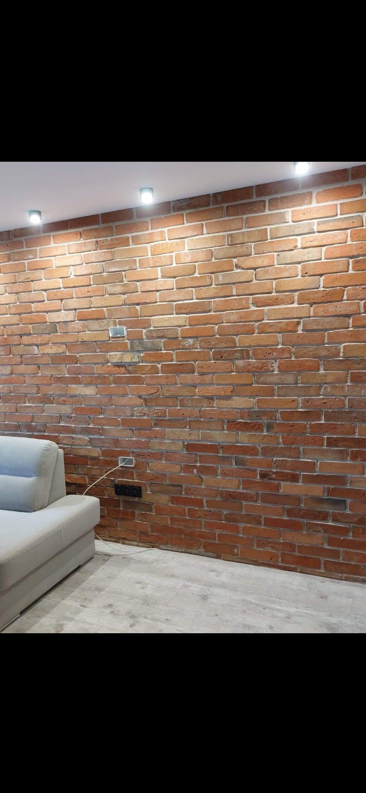 Ściana z cegły klinkierowej w salonie, oświetlona punktowymi lampami sufitowymi. Widoczny fragment sofy i gniazdka elektryczne na ścianie.