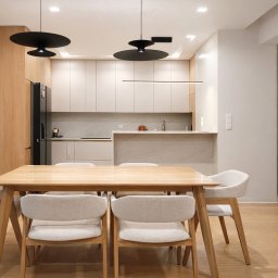 Sylwia Rybacka Architekt&Projektowanie Wnętrz - Jasna, nowoczesna jadalnia z drewnianym stołem i krzesłami na tle kuchni z beżowymi szafkami i czarną lodówką. Minimalistyczne lampy wiszące.