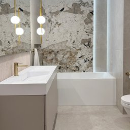 Sylwia Rybacka Architekt&Projektowanie Wnętrz - Nowoczesna łazienka z marmurową ścianą, białą wanną i umywalką z szafką w odcieniach beżu. Złote detale armatury i lamp dodają elegancji. Minimalistyczny design.
