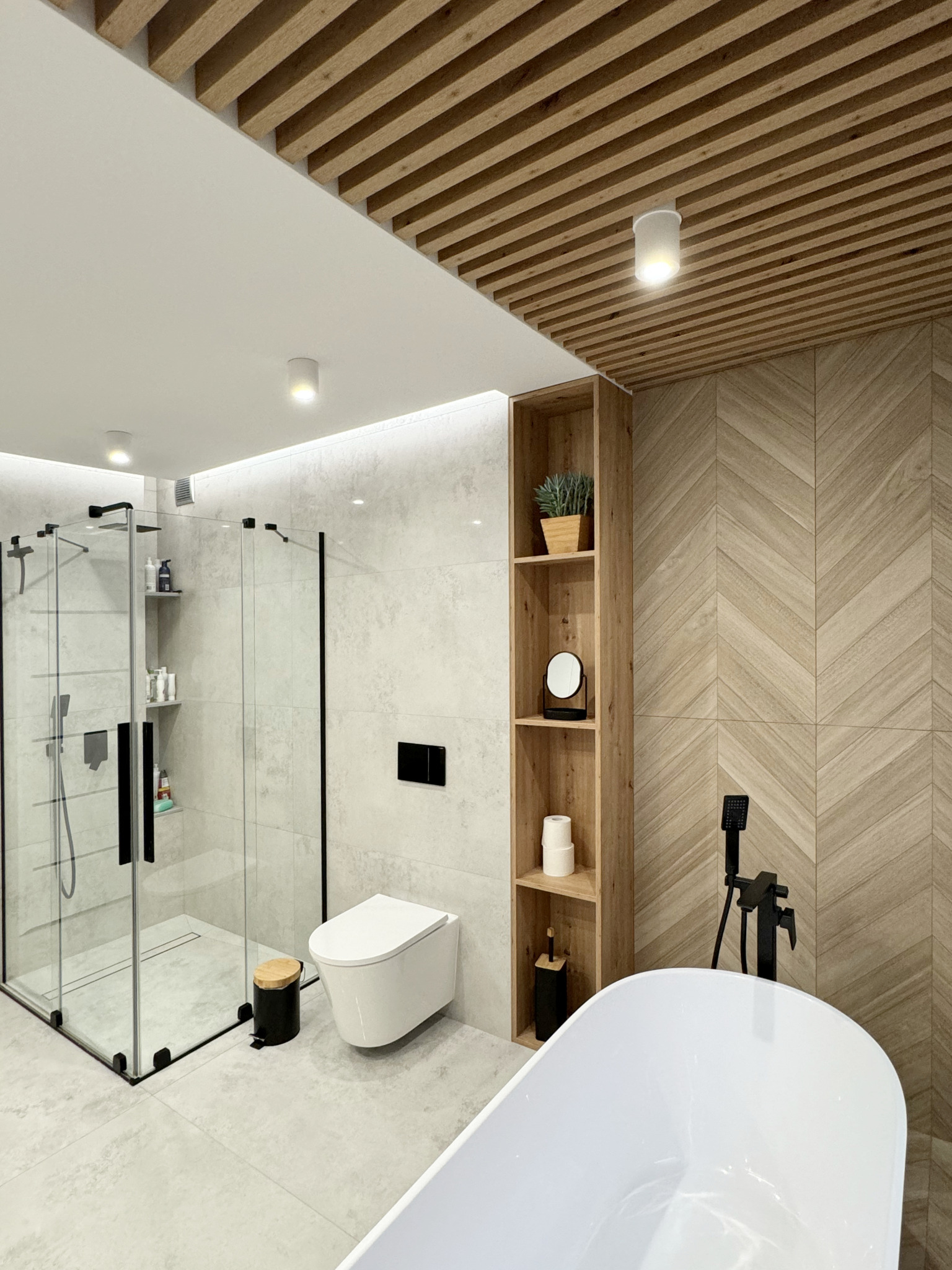 Nowoczesna łazienka z kabiną prysznicową, toaletą podwieszaną, drewnianym regałem i wanną wolnostojącą. Drewniany sufit lamelowy i płytki w jodełkę dodają ciepła.