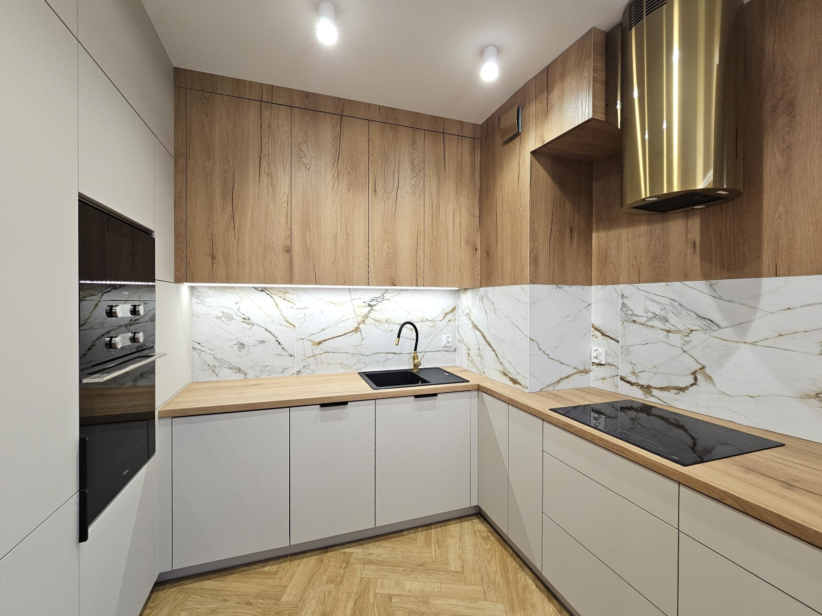 Nowoczesna kuchnia w kształcie litery L z białymi szafkami, drewnianymi blatami i złotym okapem. Marmurowe płytki na ścianie dodają elegancji.