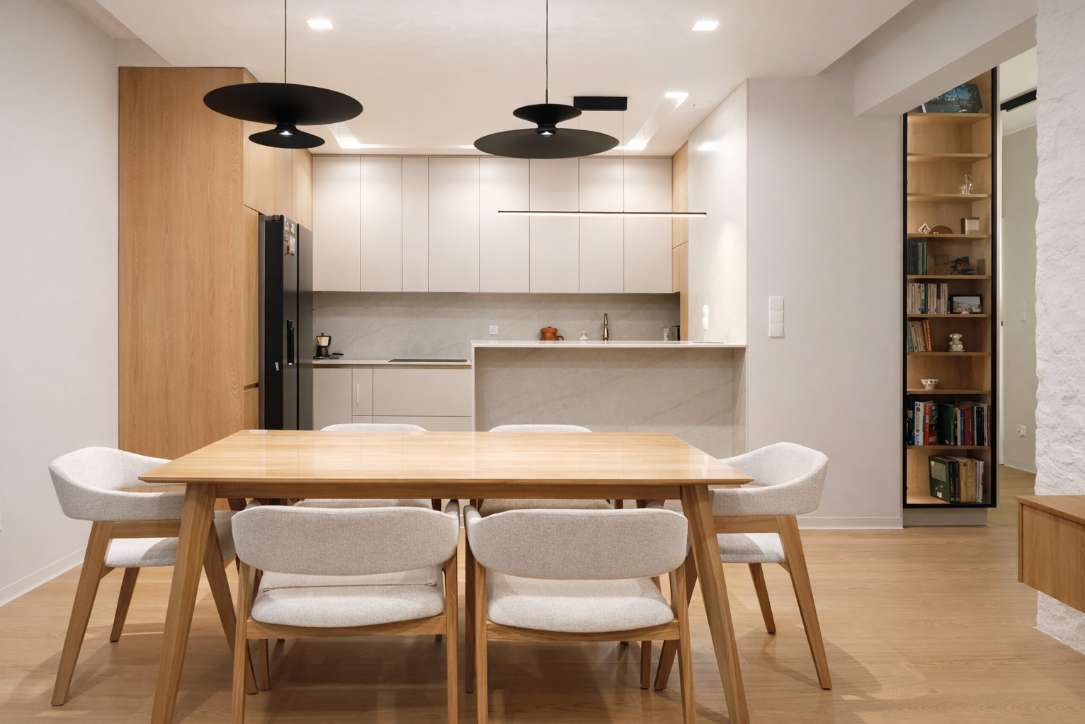 Jasna, nowoczesna jadalnia z drewnianym stołem i krzesłami na tle kuchni z beżowymi szafkami i czarną lodówką. Minimalistyczne lampy wiszące.