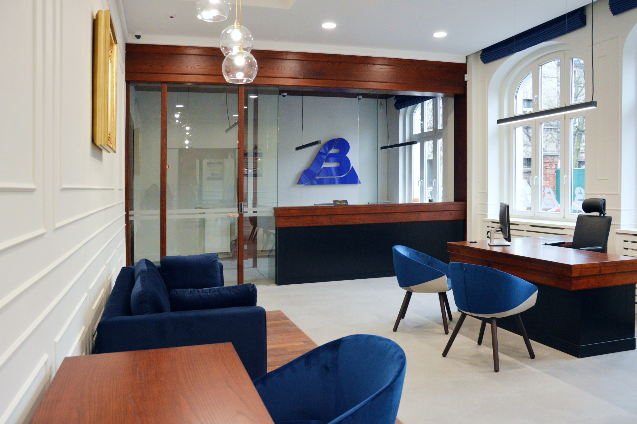 Nowoczesne biuro z recepcją, sofą i krzesłami w odcieniach granatu. Widoczne logo firmy na ścianie, drewniane elementy wykończenia i duże okno z naturalnym światłem.