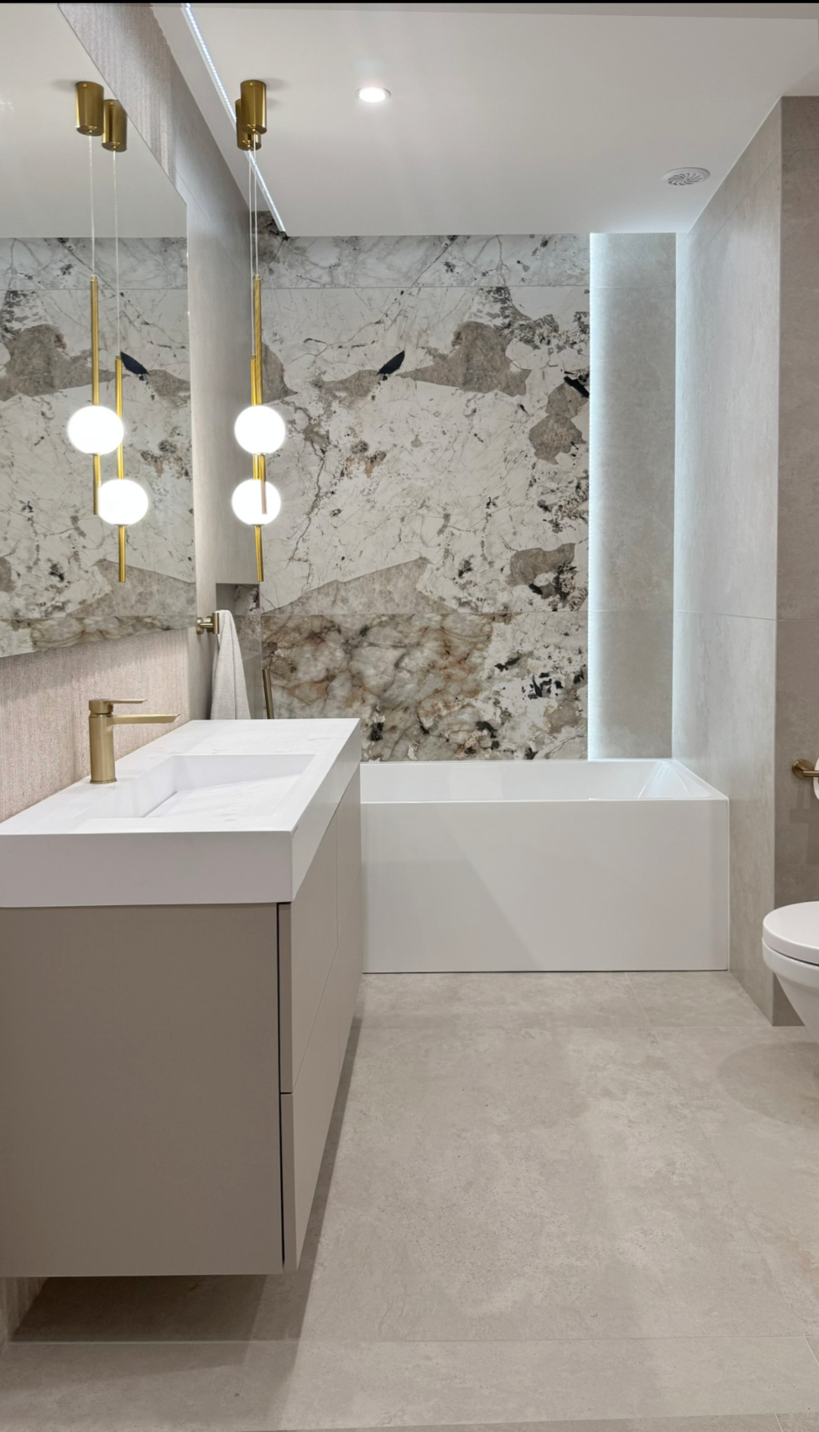 Nowoczesna łazienka z marmurową ścianą, białą wanną i umywalką z szafką w odcieniach beżu. Złote detale armatury i lamp dodają elegancji. Minimalistyczny design.