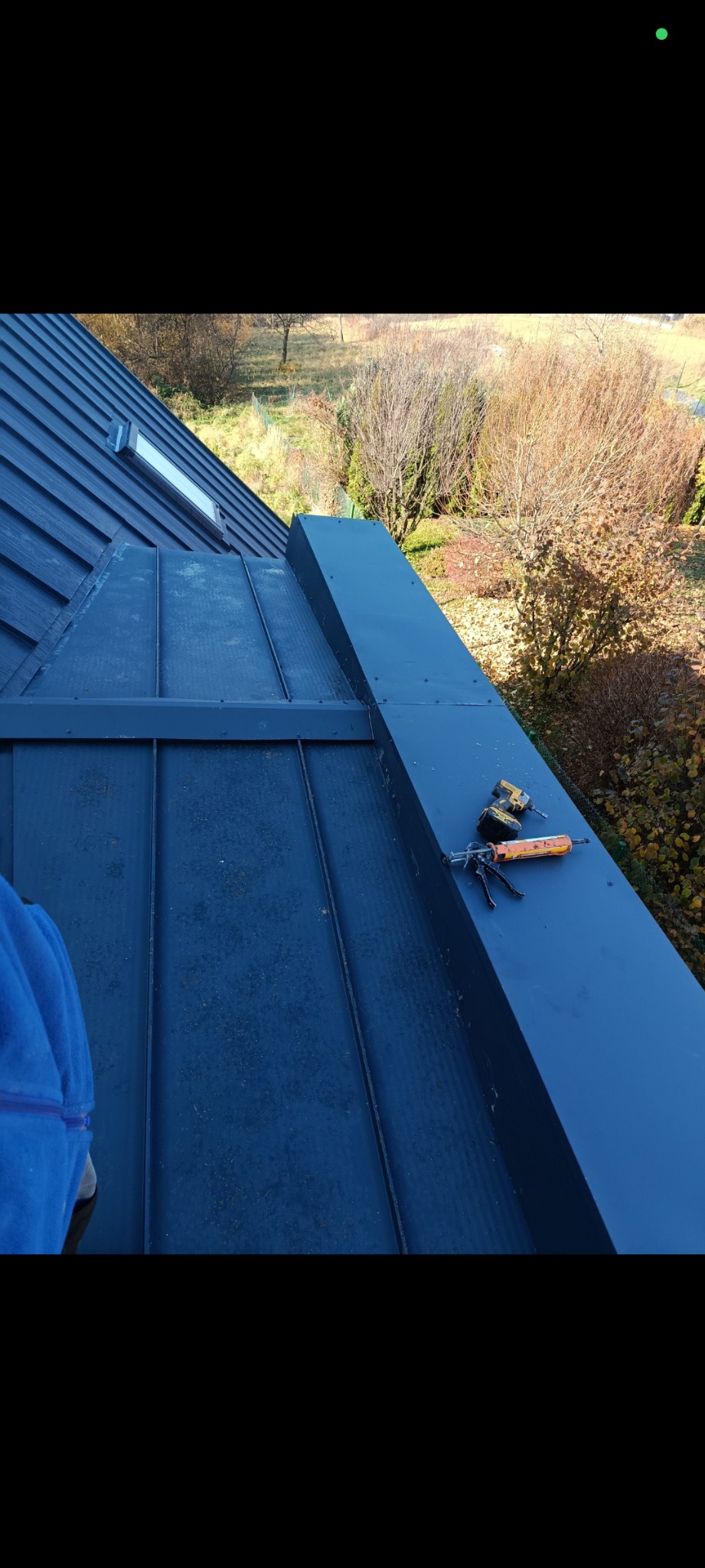 Ciemny, metalowy dach z oknem dachowym i nową obróbką blacharską. Na obróbce leżą narzędzia: wkrętarka i pistolet do silikonu. Widok z góry na jesienne drzewa.