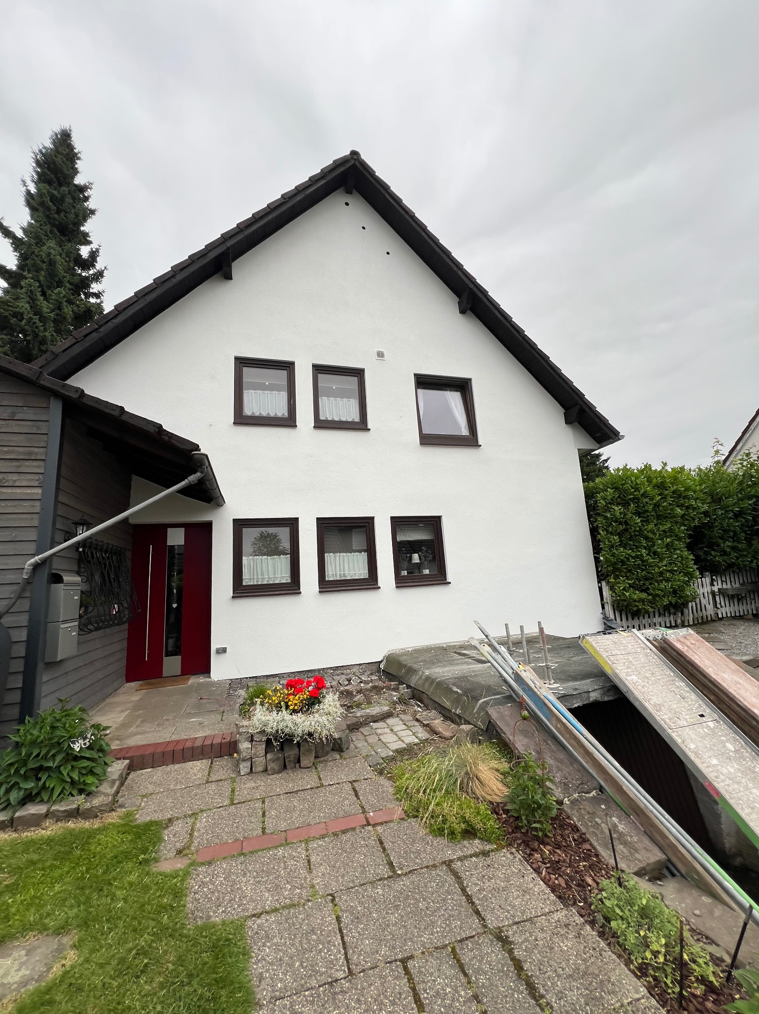 Biały dom po renowacji z brązowymi oknami i czerwoną stolarką drzwiową. Widoczne rusztowania i elementy wykończeniowe. Ujęcie z dołu, pochmurne niebo.