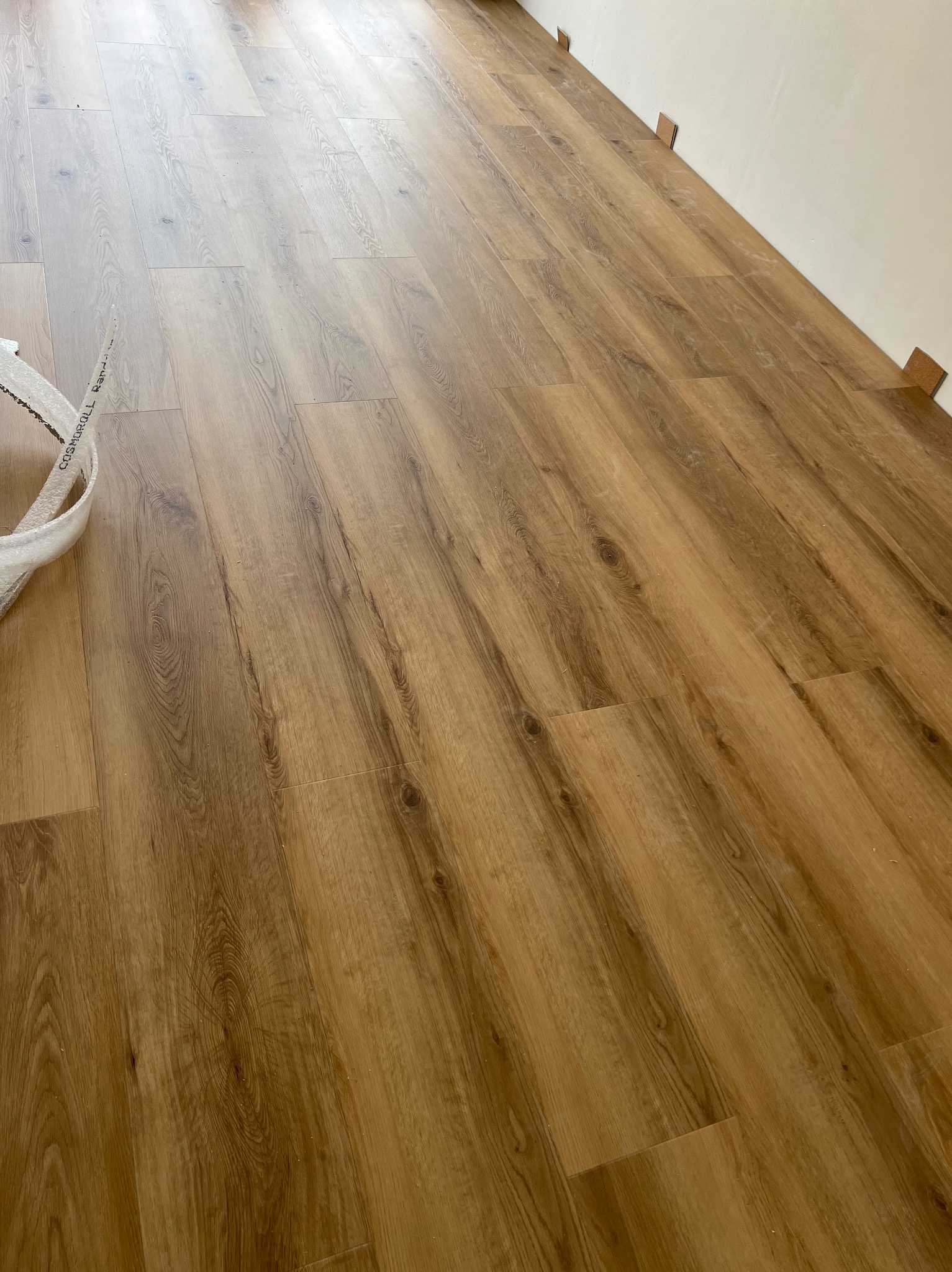 Ułożony parkiet w odcieniach brązu, widoczne listwy dystansowe przy ścianie i fragment opakowania. Podłoga z wyraźnym wzorem drewna, ułożona równolegle do ściany.