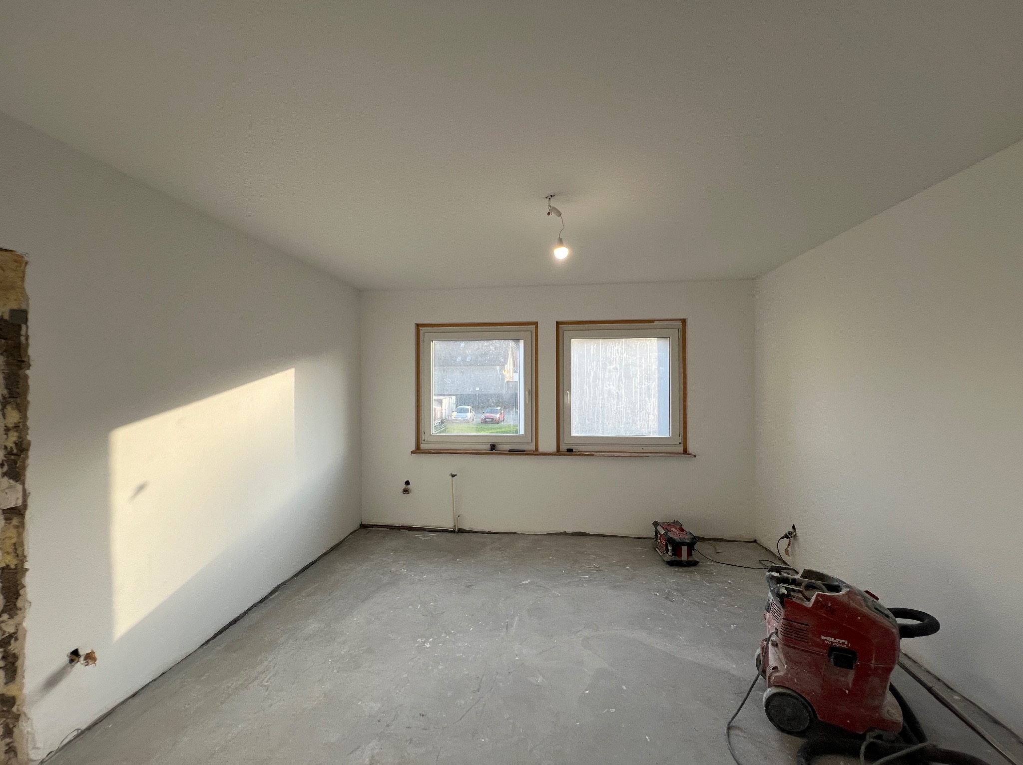 Wnętrze pokoju w trakcie remontu: świeżo pomalowane ściany na biało, surowa podłoga, dwa okna i widoczne narzędzia (odkurzacze) na podłodze. Fragment ściany bez tynku.