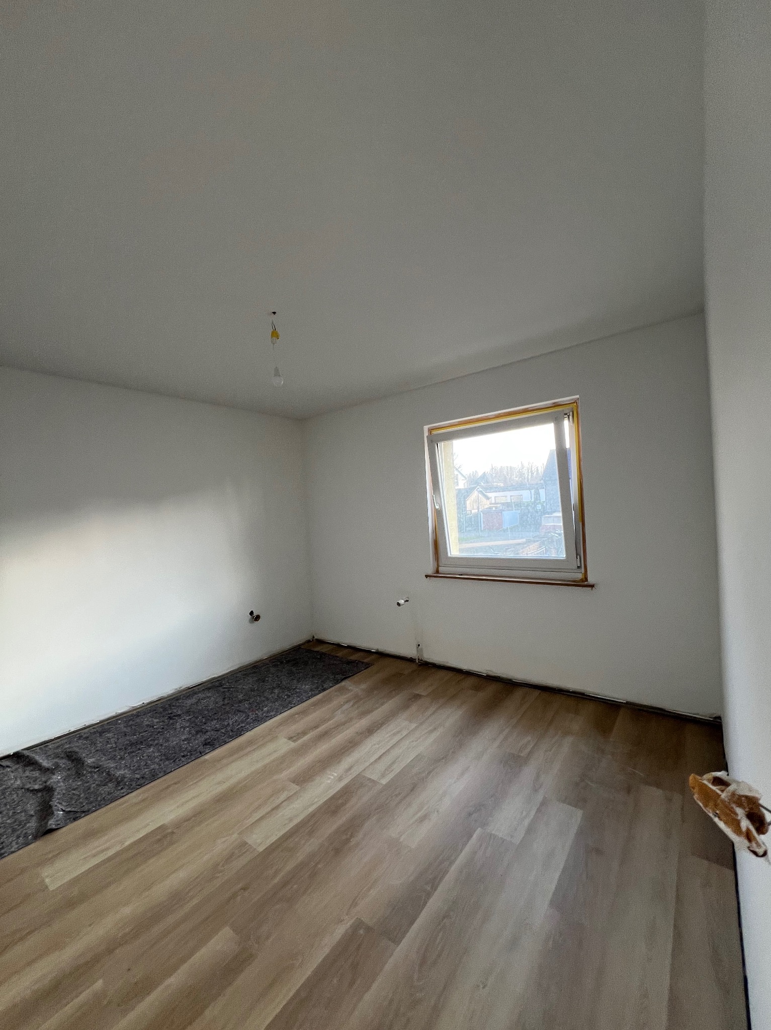 Jasne panele podłogowe w pokoju z oknem, częściowo wyłożone podkładem. Świeżo pomalowane ściany na biało, widoczne gniazdka elektryczne. Etap prac wykończeniowych.