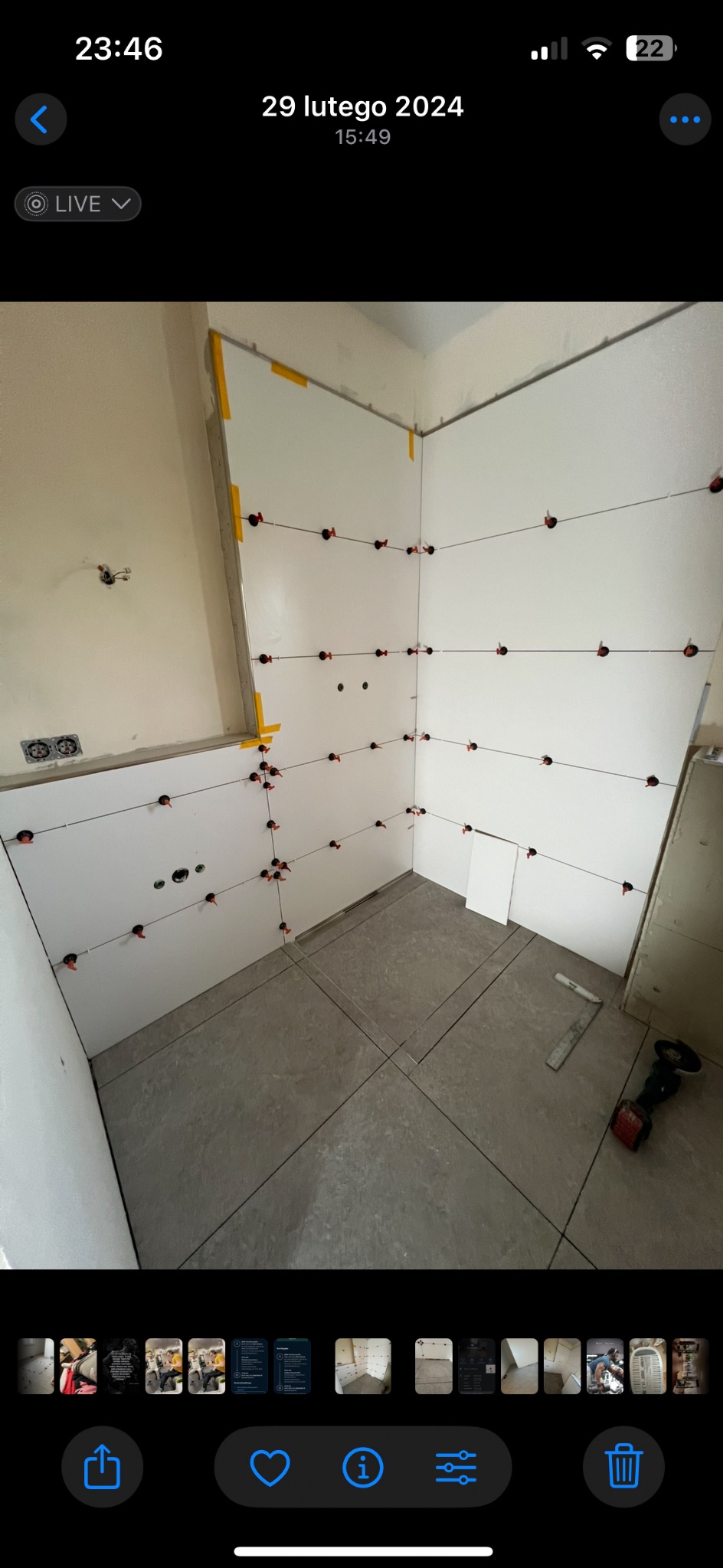 Białe płytki ścienne w trakcie montażu, z użyciem poziomujących klipsów i sznurków, na szarych płytkach podłogowych. Widoczne narzędzia i fragment instalacji elektrycznej.