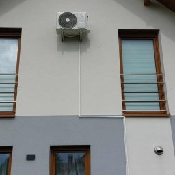 IzoKlima Zbigniew Jarmuziak - Biała jednostka zewnętrzna klimatyzacji zamontowana na elewacji domu z brązowymi oknami i szarą dolną częścią. Widoczne przewody instalacyjne.