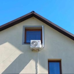 IzoKlima Zbigniew Jarmuziak - Biały klimatyzator zamontowany na elewacji domu z beżową fasadą, widoczny pod oknem z brązową ramą na tle błękitnego nieba.