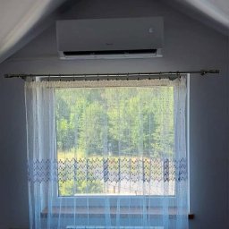 IzoKlima Zbigniew Jarmuziak - Biały klimatyzator ścienny zamontowany w pokoju na poddaszu, widoczny nad oknem z firanką i grzejnikiem. Na wyświetlaczu klimatyzatora widoczna temperatura 16 stopni.