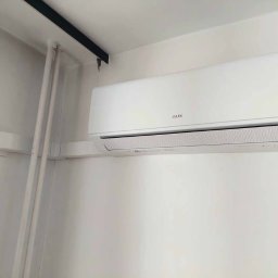 IzoKlima Zbigniew Jarmuziak - Biały klimatyzator AUX zamontowany na białej ścianie, widoczne rury instalacyjne. Minimalistyczny design, oświetlenie dzienne.