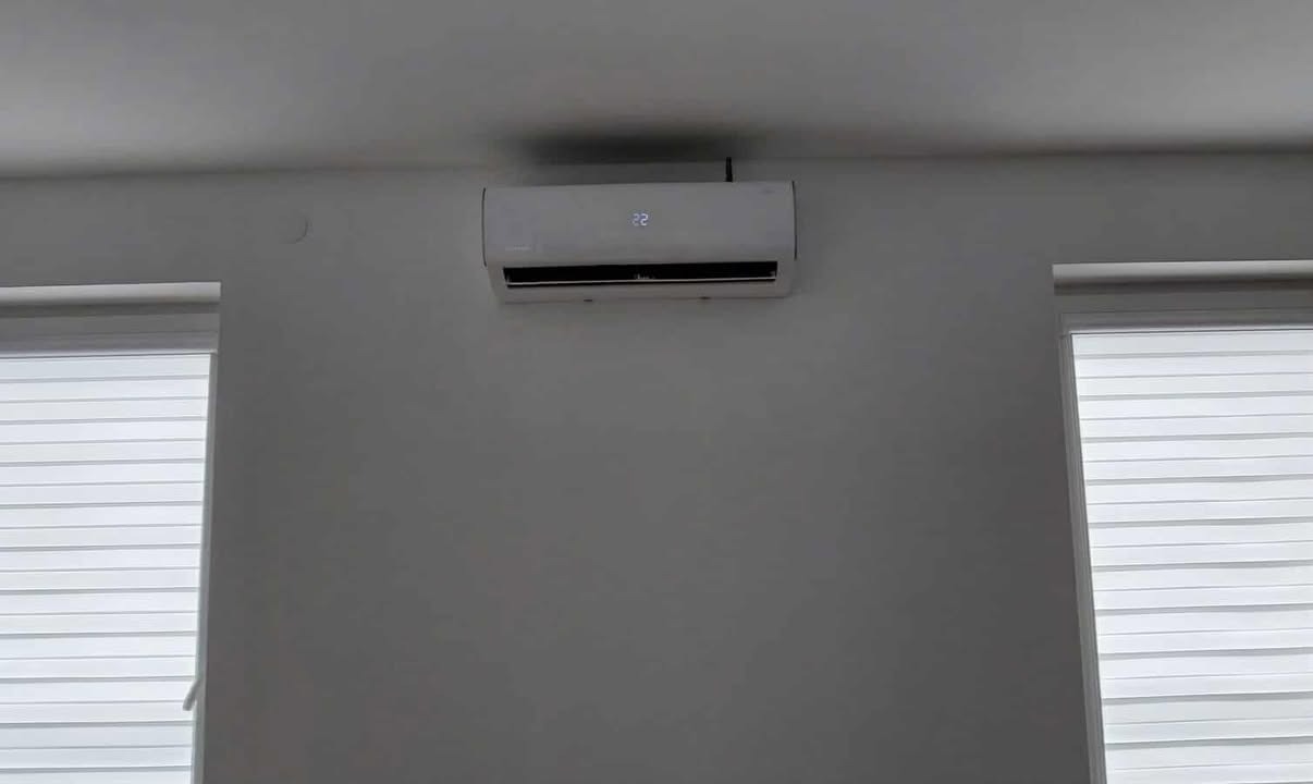 Biały klimatyzator ścienny z wyświetlaczem temperatury 22 stopnie, zamontowany w jasnym pomieszczeniu z dwoma oknami z żaluzjami poziomymi.
