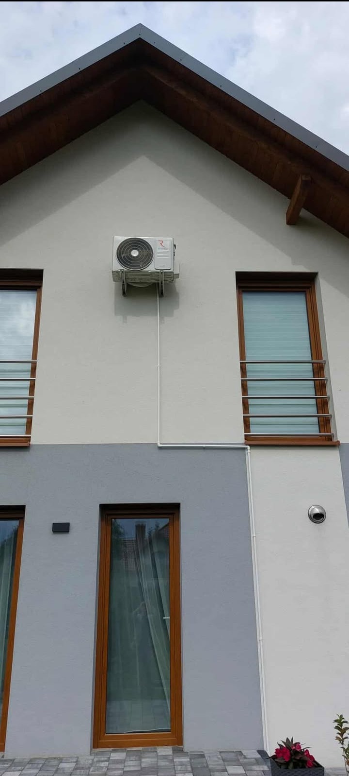 Biała jednostka zewnętrzna klimatyzacji zamontowana na elewacji domu z brązowymi oknami i szarą dolną częścią. Widoczne przewody instalacyjne.