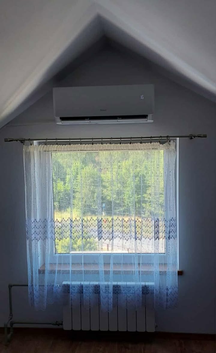 Biały klimatyzator ścienny zamontowany w pokoju na poddaszu, widoczny nad oknem z firanką i grzejnikiem. Na wyświetlaczu klimatyzatora widoczna temperatura 16 stopni.