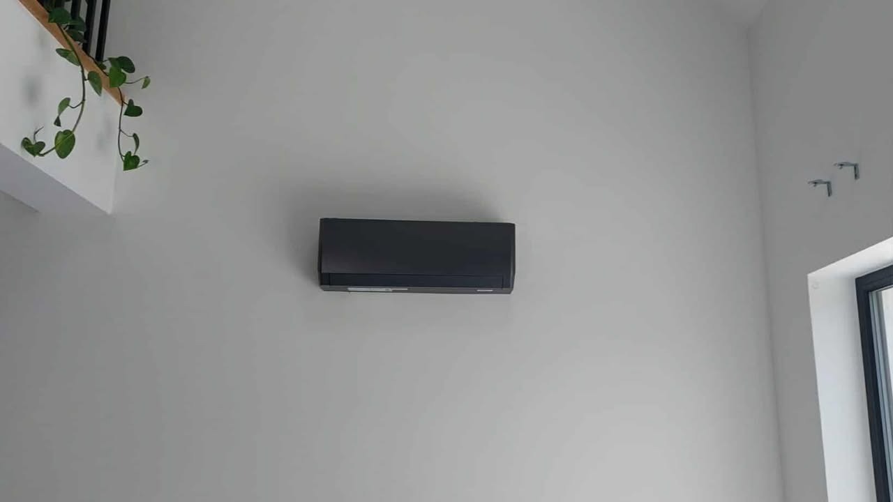 Czarny klimatyzator ścienny w jasnym wnętrzu, z rośliną zwisającą z góry i oknem po prawej stronie. Minimalistyczny design, nowoczesne wykończenie.