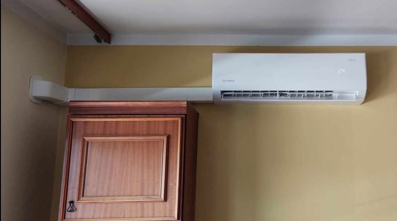 Biały klimatyzator Rotenso zamontowany na ścianie nad drewnianą szafką, widoczne przewody instalacyjne w białej listwie. Ściana w odcieniach beżu.