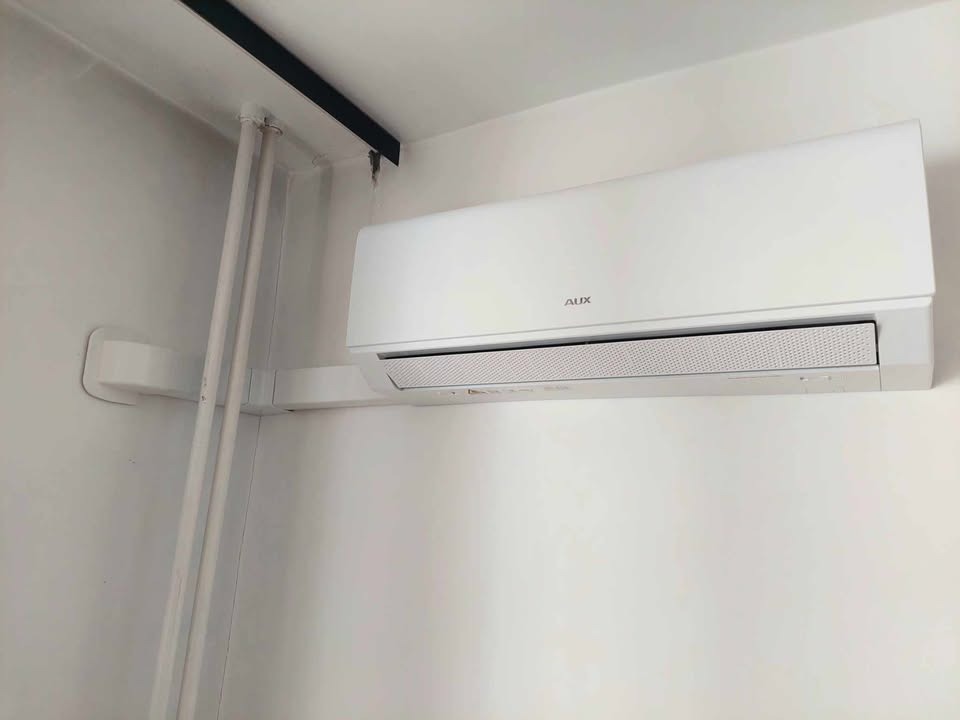 Biały klimatyzator AUX zamontowany na białej ścianie, widoczne rury instalacyjne. Minimalistyczny design, oświetlenie dzienne.