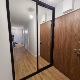 MULTISTOL Marcin Juszczak - Nowoczesna szafa wnękowa z lustrzanymi drzwiami przesuwnymi w przedpokoju. Czarna rama, drewnopodobne drzwi wejściowe, jasne ściany i podłoga.