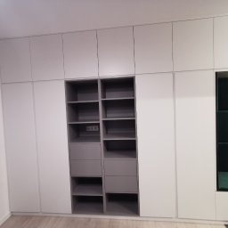 MULTISTOL Marcin Juszczak - Biało-szara szafa wnękowa z półkami, szufladami i zielonym siedziskiem w przedpokoju. Widoczne gniazdka elektryczne w jednej z półek. Nowoczesny design.