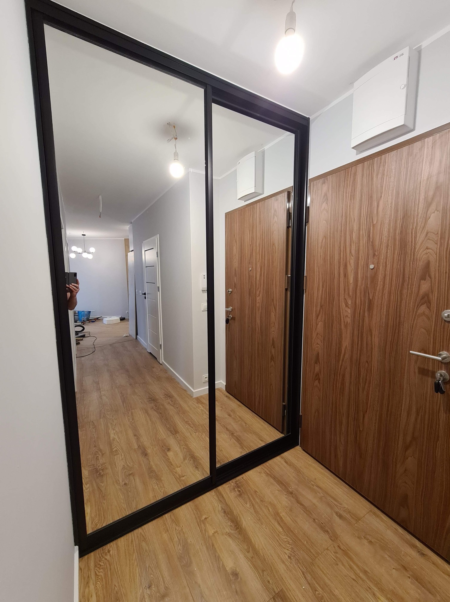 Nowoczesna szafa wnękowa z lustrzanymi drzwiami przesuwnymi w przedpokoju. Czarna rama, drewnopodobne drzwi wejściowe, jasne ściany i podłoga.
