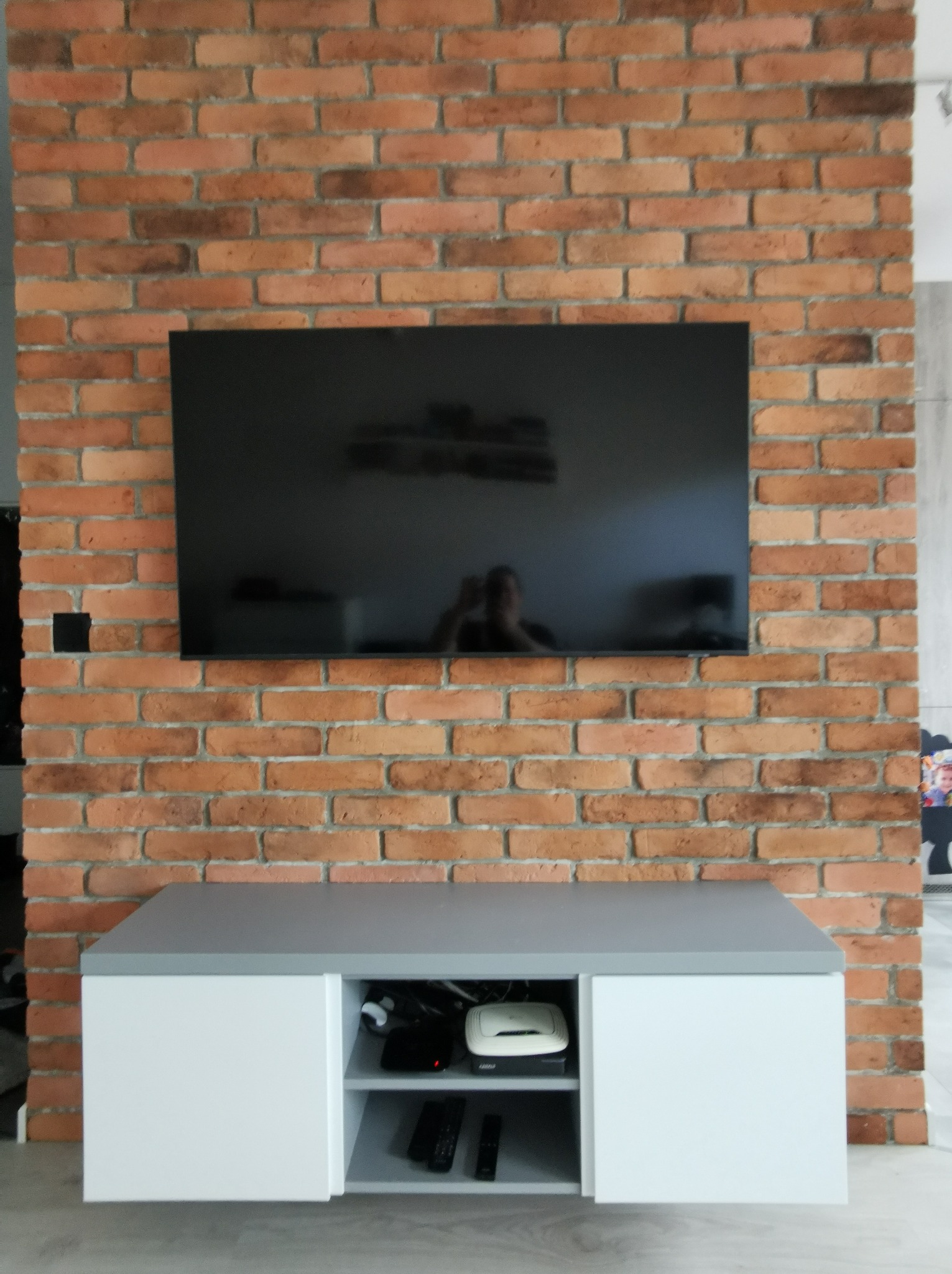 Telewizor na ceglanej ścianie nad białą szafką RTV z szarym blatem. Widoczne kable i sprzęt wewnątrz szafki. Minimalistyczny design.