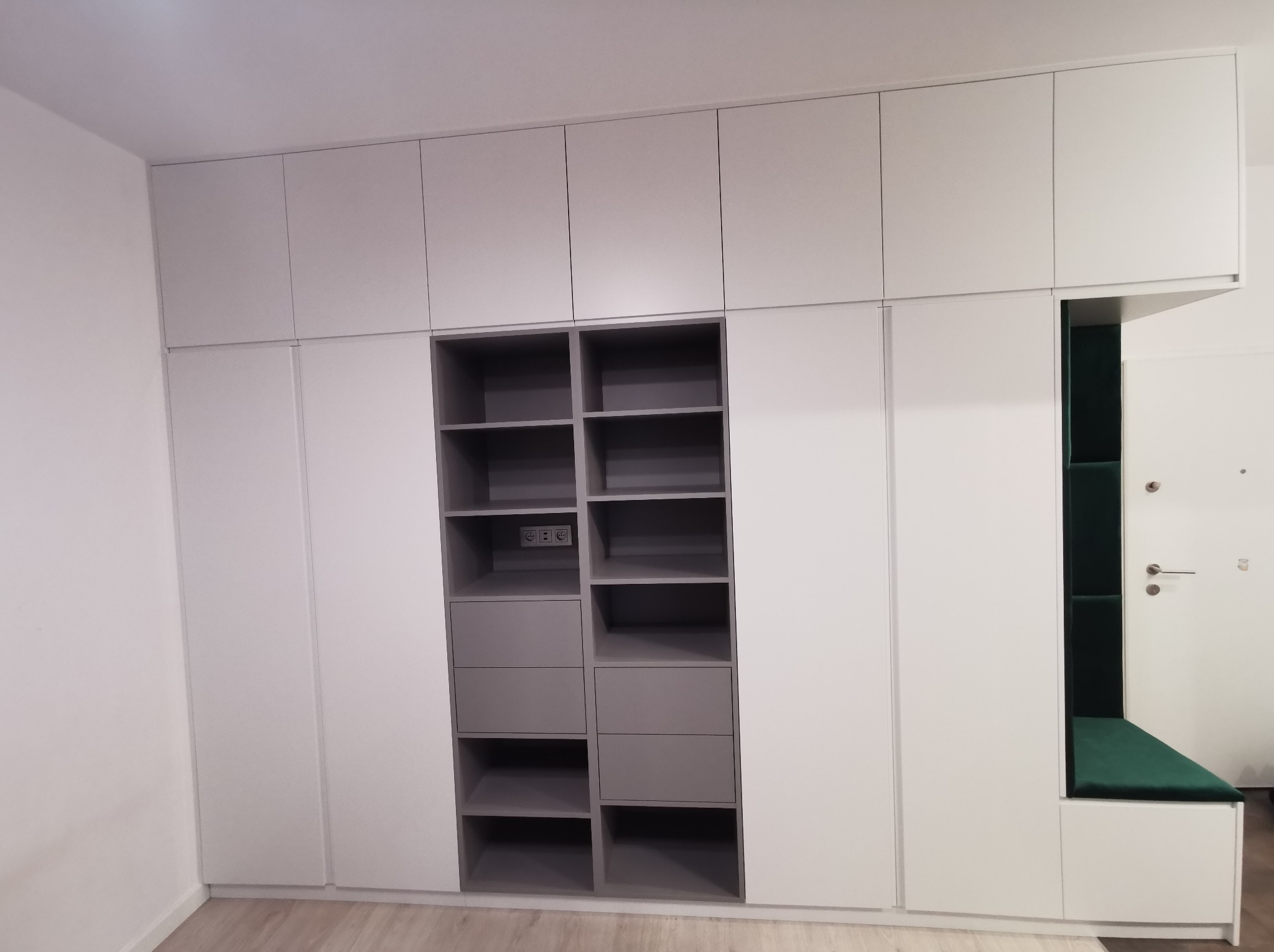 Biało-szara szafa wnękowa z półkami, szufladami i zielonym siedziskiem w przedpokoju. Widoczne gniazdka elektryczne w jednej z półek. Nowoczesny design.