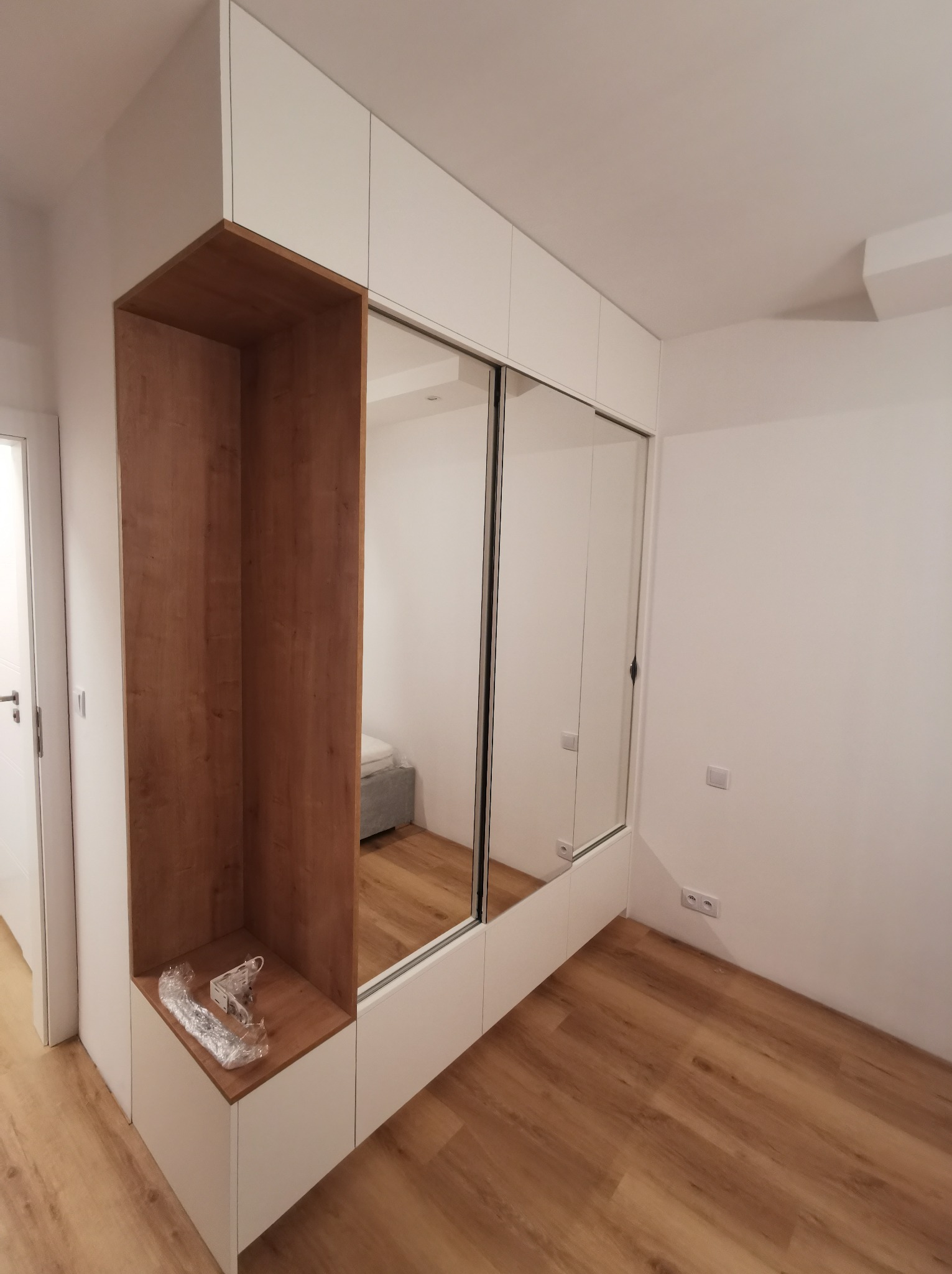 Biała szafa wnękowa z lustrzanymi drzwiami przesuwnymi i drewnianą wnęką, widoczna na tle parkietu i białych ścian. W wnęce leżą akcesoria montażowe.
