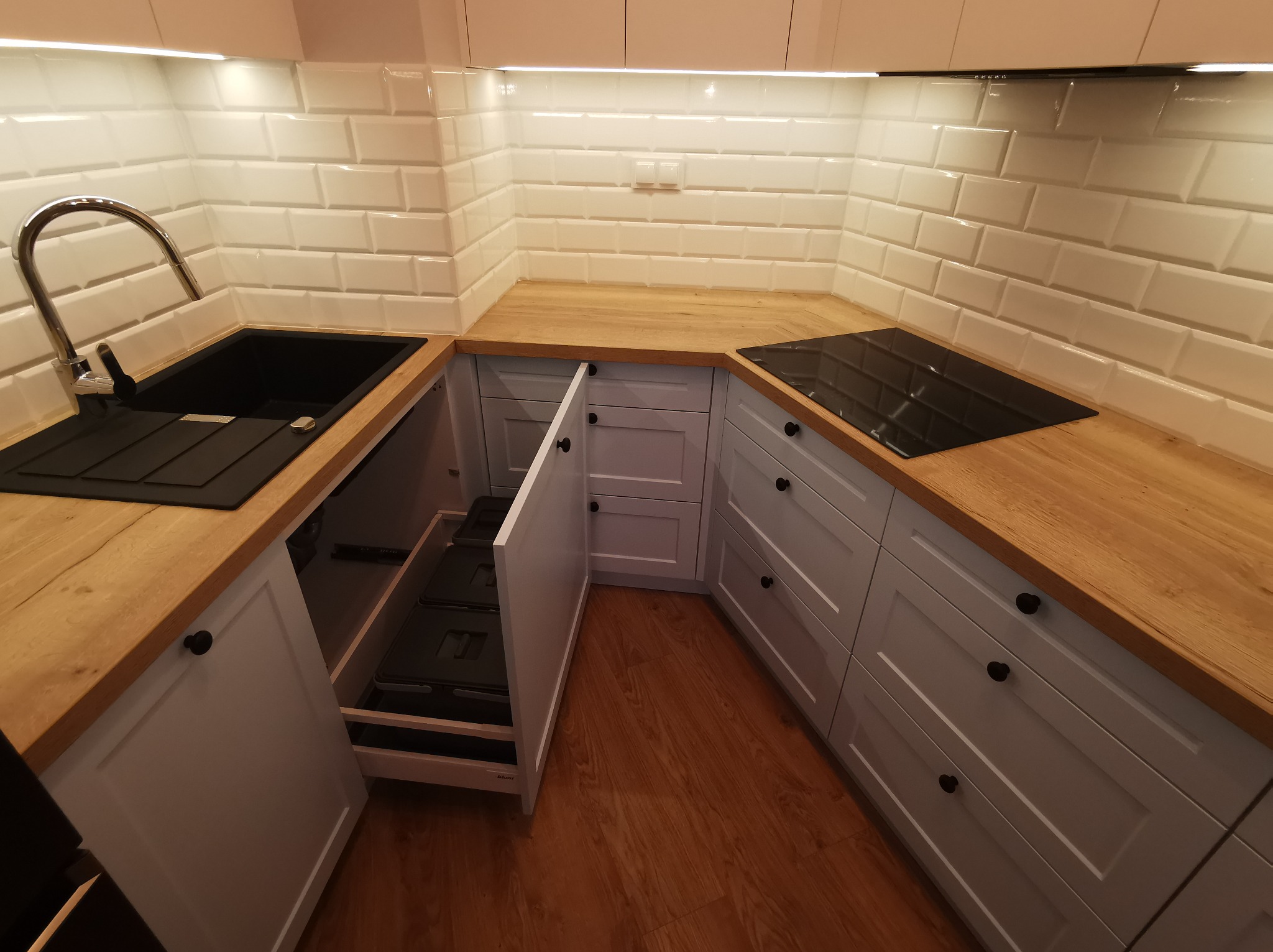 Jasna kuchnia z drewnianym blatem, białymi płytkami i otwartą szafką z wysuwanymi koszami. Nowoczesny design z czarnym zlewem i płytą indukcyjną.