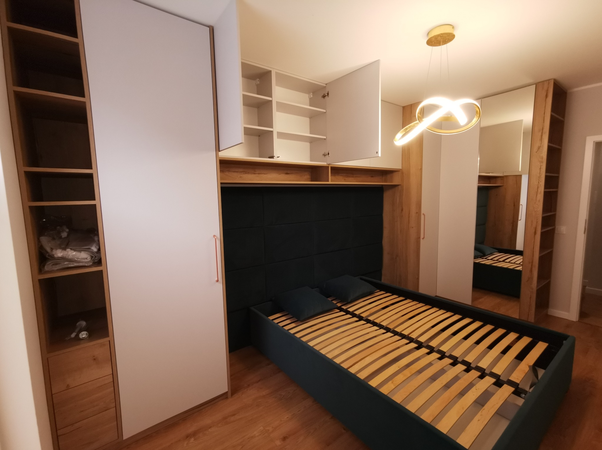 Nowoczesna sypialnia z zabudową meblową: szafy z białymi frontami, drewniane regały, łóżko z tapicerowanym zagłówkiem i designerska lampa. Całość w ciepłej tonacji.