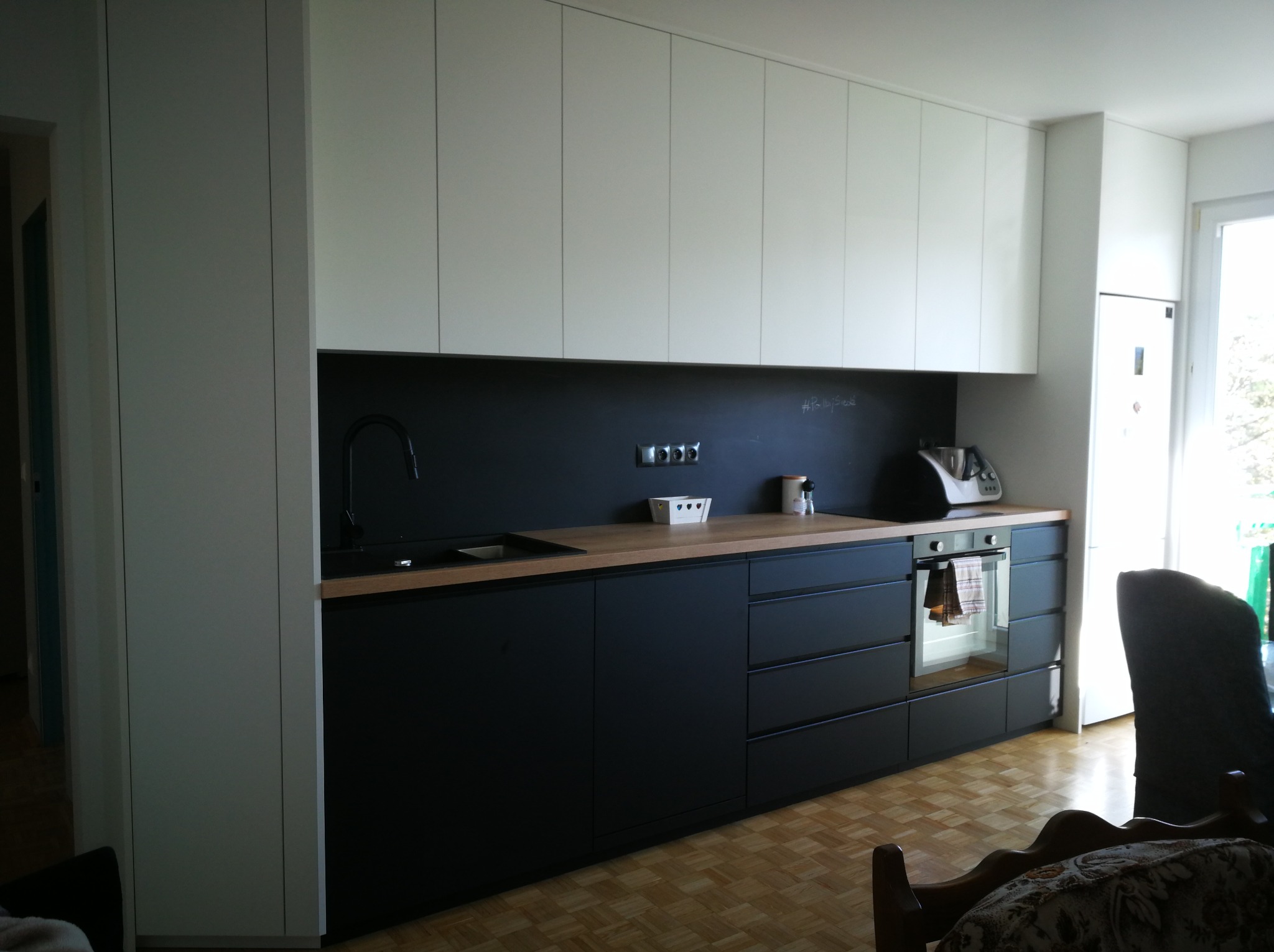 Nowoczesna kuchnia z czarnymi szafkami dolnymi i białymi górnymi, drewniany blat, minimalistyczny design, widok na okno i fragment salonu.