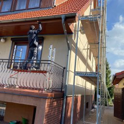 Kamil Wojtala - Pracownik w czarnej kurtce maluje brązowe podbitki dachowe domu z czerwonym dachem, stojąc na drabinie na balkonie z kutą balustradą. Obok elewacji widoczne jest rusztowanie.