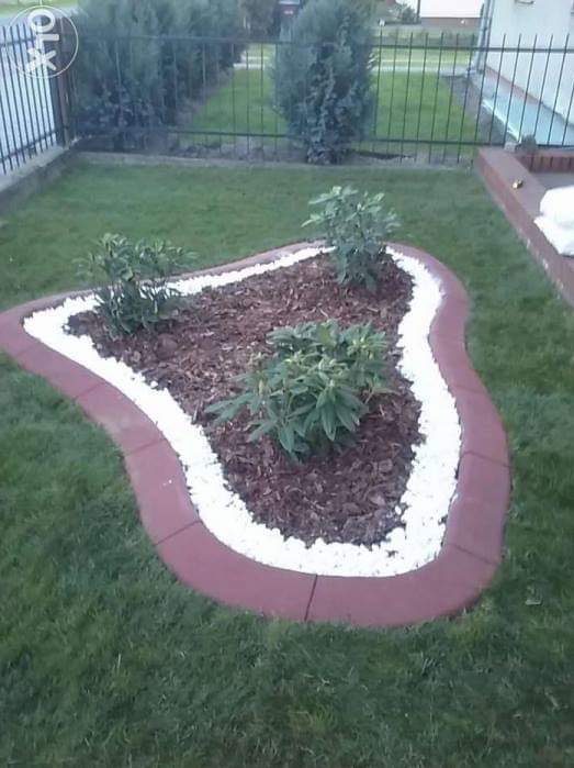 Kształtna rabata z korą, obwódką z białych kamieni i ceglanym obrzeżem na trawniku. Widok z góry na fragment ogrodu z niską, metalową siatką w tle.