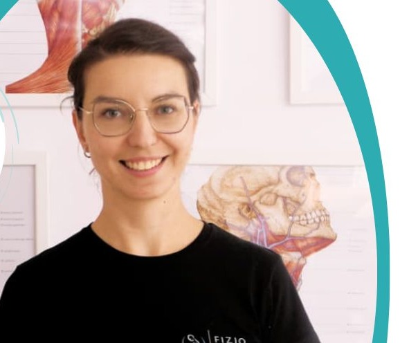 Uśmiechnięta kobieta w okularach, czarnej koszulce z logo, na tle ściany z grafikami anatomicznymi. Portret fizjoterapeutki w gabinecie.