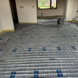 Usługi sanitarno-grzewcze Stanisław Parkoła - Ułożona instalacja ogrzewania podłogowego w surowym wnętrzu domu, widoczne rury na folii z rastrem, przygotowane do zalania posadzką. W tle okno i fragment instalacji.
