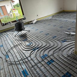 Usługi sanitarno-grzewcze Stanisław Parkoła - Ułożona instalacja ogrzewania podłogowego na folii z nadrukiem, widoczna cewka z rurą i okno z widokiem na dom. Etap przed wylewką.