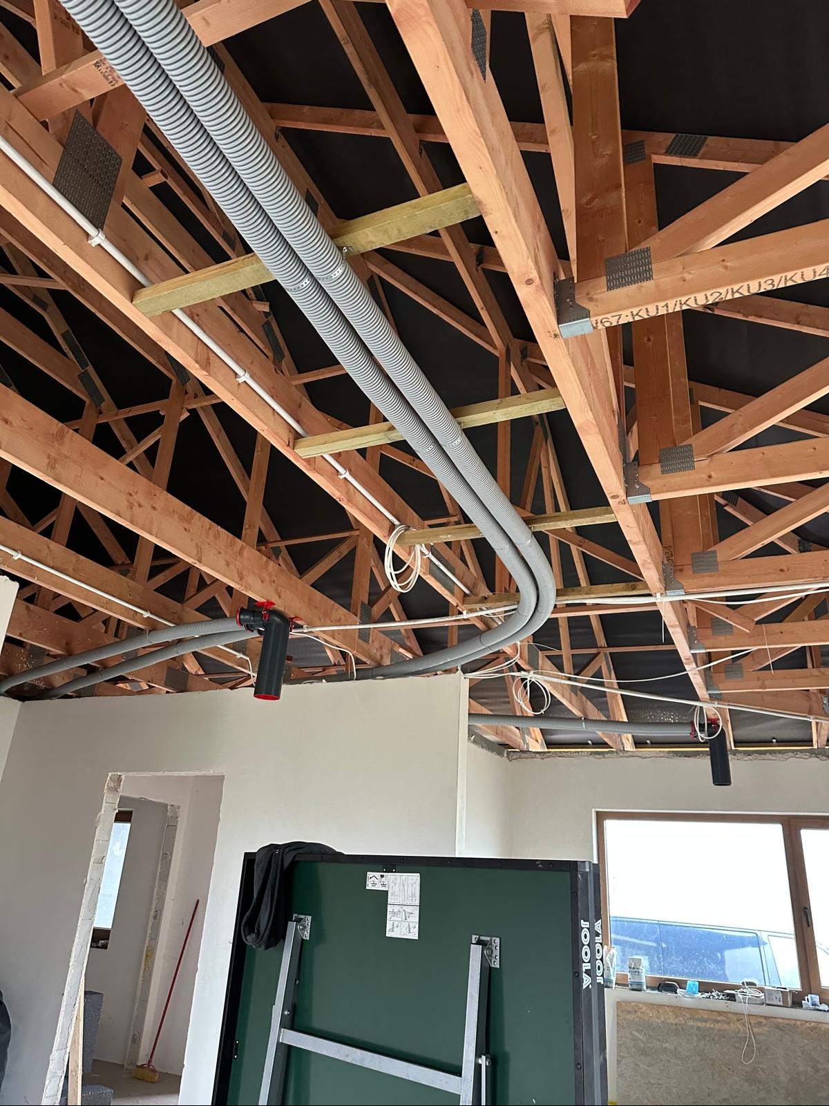 Szare, karbowane rury wentylacyjne pod drewnianą konstrukcją dachu, widoczne elementy instalacji elektrycznej i fragment stołu do ping-ponga. Prace instalacyjne w surowym wnętrzu.